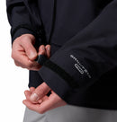A person adjusts the Velcro cuff strap on a dark Columbia Mens Saudan Pro 3L Waterproof Shell Jacket, with the logo visible, while holding the waterproof sleeve steady.