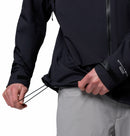 A person wearing Columbias Mens Saudan Pro 3L Waterproof Shell Jacket, with Omni-Tech on the sleeve and paired with light gray pants, adjusts the hem drawcord. Only their torso and hands are visible.