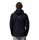 A person wearing a black Columbia Mens Saudan Pro 3L Waterproof Shell Jacket with the logo on the back is shown facing away from the camera, paired with light gray pants.
