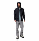 A man wearing a Columbia Mens Saudan Pro 3L Waterproof Shell Jacket in black, a light blue shirt, gray pants, and gray hiking shoes stands facing forward against a white background.
