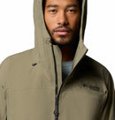 A person wears a partially zipped, hooded Men’s Saudan Pro 3L Waterproof Shell Jacket by Columbia in light olive, with the Columbia Titanium logo visible on the chest.