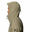 A person in profile wears the Columbia Mens Saudan Pro 3L Waterproof Shell Jacket in tan, with black logos on the chest and sleeve. The hood is up and covers most of the head, making it ideal for extreme conditions.