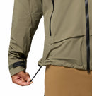 A person in the Columbia Mens Saudan Pro 3L Waterproof Shell Jacket and tan pants pulls the adjustable drawcord at the hem. The image highlights their hand, jacket hem, and detailed zipper features.