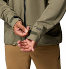 A person adjusts the Velcro cuff strap on a Columbia Mens Saudan Pro 3L Waterproof Shell Jacket in olive green, featuring Omni-Tech Pro on the sleeve, paired with tan pants. Only the torso and hands are visible.
