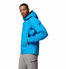A man wearing a Columbia Mens Saudan Pro 3L Waterproof Shell Jacket in bright blue and light gray pants stands with one hand in his pocket, posing against a plain white background.