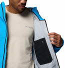 A person wearing a light beige Columbia t-shirt and a blue Columbia Mens Saudan Pro 3L Waterproof Shell Jacket holds open the jacket to reveal an inner zippered pocket on the light gray lining.