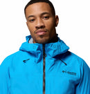 A man wears a bright blue Columbia Mens Saudan Pro 3L Waterproof Shell Jacket, looking at the camera with a neutral expression. The Columbia logo is visible on the left chest.