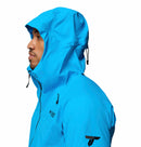 A person wearing a bright blue Columbia Mens Saudan Pro 3L Waterproof Shell Jacket is shown from the side with the hood up. Black drawstrings and Columbia logos appear on the chest and sleeve, set against a white background.