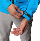 A person adjusts the Velcro cuff of a bright blue Columbia Mens Saudan Pro 3L Waterproof Shell Jacket sleeve with one hand; they are wearing light gray pants. The Omni-Tech Pro logos are visible on the jacket.
