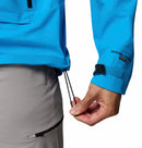 A person adjusts the hem drawstring of a Columbia Mens Saudan Pro 3L Waterproof Shell Jacket. Only their lower torso and hand are visible, and they wear light gray pants with a zippered pocket.
