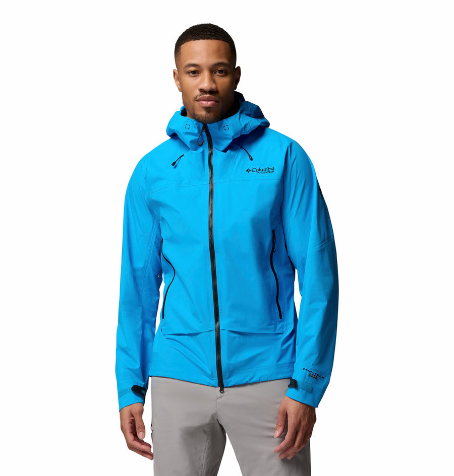 A man faces forward against a plain white background, wearing the Columbia Mens Saudan Pro 3L Waterproof Shell Jacket in bright blue with a front zipper, paired with light gray pants.