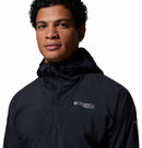 A person with dark curly hair wears a black Columbia Mens Khyex Pro™ Wind Jacket, featuring a hood and partially zipped front. The Columbia logo appears on the left chest. The background is plain white.