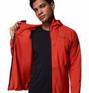 A person wears a red Columbia Mens Khyex Pro™ Wind Jacket over a black shirt, standing against a white background and holding one side open to reveal the interior and highlight its UPF 50 protection.