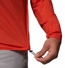 A person tightens the adjustable drawstring at the hem of a bright red Columbia Mens Khyex Pro™ Wind Jacket, which is ultralight and offers UPF 50 protection, while wearing light-colored pants.