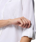 A woman wearing the Columbia Womens Silver Ridge Utility II Long-Sleeve Shirt in white is shown rolling up and buttoning her left sleeve, with only her torso and arms visible.