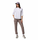 A woman stands against a white background wearing the Columbia Womens Silver Ridge Utility II Long-Sleeve Shirt in white, taupe cargo pants, a light purple baseball cap, and beige sneakers. One hand rests in her pocket as she looks to the side.