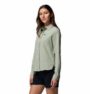 A woman with long brown hair stands against a white background, smiling gently while wearing the Columbia Womens Silver Ridge Utility II Long-Sleeve Shirt in light green and black shorts, her arms relaxed at her sides.