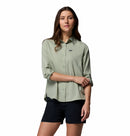 A woman stands against a white background wearing the Columbia Womens Silver Ridge Utility II Long-Sleeve Shirt in light green and black shorts, sleeves rolled up, looking at the camera with a neutral expression.