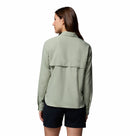 A woman with straight brown hair stands with her back to the camera, wearing a Columbia Womens Silver Ridge Utility II Long-Sleeve Shirt in light green and black shorts against a plain white background.