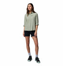 A woman stands against a white background wearing the Columbia Womens Silver Ridge Utility II Long-Sleeve Shirt in light green, paired with black shorts, gray socks, and black sneakers. Her sleeves are rolled up and she looks slightly to the side with a relaxed expression.