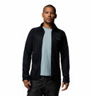 A man faces forward against a plain white background wearing a light blue t-shirt, black pants, and the Columbia Mens Summit Step™ Full Zip Technical Fleece jacket in black with a lightweight design.