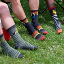 Three people’s lower legs and feet are visible among pink petals on grass, wearing colorful, mismatched Darn Tough Mens Sunset Ridge Micro Crew Lightweight Hiking Socks with animal, stripe, and bird patterns.