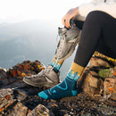 At sunrise, a person sits on a rocky ledge in black leggings and colorful Darn Tough Women’s Northwoods Micro Crew Midweight Hiking Socks, featuring tree designs. One hiking shoe is off—showcasing the Vermont-made sock—while the other shoe remains on. Mountains stretch across the background.