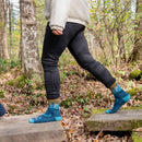 A person wearing black pants, a cream-colored jacket, and Darn Tough Women’s Northwoods Micro Crew Midweight Hiking Socks in a blue pattern—made in Vermont—walks on wooden planks through a forest with fallen leaves and trees.