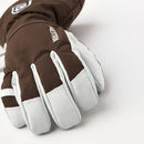 A close-up of a brown and white Hestra Army Leather Heli Ski Glove from the Alpine Pro collection, featuring the Hestra brand name on the back, displayed resting on a white surface.