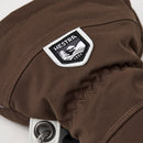 Close-up of the Hestra Army Leather Heli Ski Glove in brown, displaying a white-and-black Hestra 1936 patch with an image of two hands. The durable fabric, detailed stitching, and metal eyelet at the cuff reflect the superior craftsmanship of Hestra’s Alpine Pro collection.