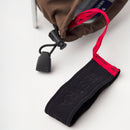 A close-up of a brown bag with a red and black Hestra-branded strap, reminiscent of the iconic design featured on the Army Leather Heli Ski Glove by Hestra, shows a plastic cord lock and drawstring resting on a white surface.