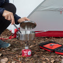 An individual cooks with an MSR Alpine StowAway Pot on a portable camping stove outside their tent, using this durable cookware designed for outdoor adventures. The ground is strewn with dry leaves, and a red utensil case lies nearby.