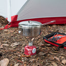 An MSR Alpine StowAway Pot heats on a small gas stove next to a red and white tent, while nearby, a black and red case holds durable MSR cookware and camping utensils on the ground covered with dry leaves.