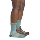 A person stands sideways wearing one hiking boot and one Darn Tough Womens Mountaineering Micro Crew Heavyweight Hiking Sock in light blue-green with teal accents, showing a comparison between the sock and the boot.