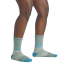 A person stands on a white background, showing only the lower legs and feet, wearing Darn Tough Womens Mountaineering Micro Crew Heavyweight Hiking Socks in light green with blue accents.