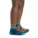 A person wearing brown hiking boots and Darn Tough Women’s Northwoods Micro Crew Midweight Hiking Socks—colorful merino wool socks with tree patterns, made in Vermont—stands on one foot; the other foot, clad only in a sock, rests sideways against the standing ankle.