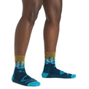 A person wearing Darn Tough Women’s Northwoods Micro Crew Midweight Hiking Socks, featuring a blue and teal landscape design with trees and mountains, stands on a white background.