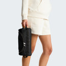 Dressed in a cream-colored hoodie and shorts, a person holds The North Face Base Camp Travel Washbag - Large by its handle, standing against a plain light background.