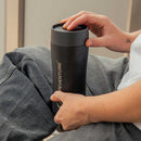 A person holds a sleek black Lifeventure One-Touch Thermal Mug with “PREVENTURE” written on it, resting it on their lap. One hand is on the stainless steel one-touch lid, and a small tattoo is visible on their wrist.