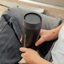 A person sits cross-legged in gray jeans and a white shirt, showing a small leaf tattoo on their left wrist, while holding a black Lifeventure One-Touch Thermal Mug featuring a convenient One-Touch Lid.