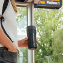 A person holding a Lifeventure One-Touch Thermal Mug in black at a train station, with a digital sign overhead and trees visible through large windows.