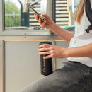 A person sitting by a sunlit window, holding a phone in one hand and a black Lifeventure One-Touch Thermal Mug in the other.