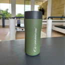 A green Lifeventure One-Touch Thermal Mug with a gray lid rests on a gym bench, surrounded by exercise equipment and large windows in the background.