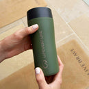 A person holding a green Lifeventure One-Touch Thermal Mug above tan mats on a tiled floor.