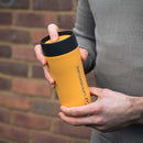 A person holding a yellow Lifeventure One-Touch Thermal Mug with a black lid stands against a brick wall, dressed in a gray long-sleeve shirt.