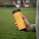 A person holds a yellow Lifeventure One-Touch Thermal Mug outdoors. In the background, there is a grassy field and some blurred houses. The individual is dressed in a light gray long-sleeve shirt, and the mug features a convenient One-Touch Lid for easy sipping on the go.