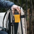 Outdoors, a person places a yellow Lifeventure One-Touch Thermal Mug into the side pocket of a beige and black backpack.