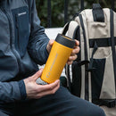 A person in a dark jacket holds the bright yellow Lifeventure One-Touch Thermal Mug with a convenient One-Touch Lid next to a beige and black backpack outdoors.