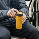 A person wearing a gray jacket holds a Lifeventure One-Touch Thermal Mug with a convenient one-touch lid while sitting on a bench, with a backpack nearby.