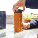 A person in a blue sweater twists the black lid of an orange Lifeventure One-Touch Thermal Mug on a sunlit table. A blue mug and a small green plant are in the blurred background.